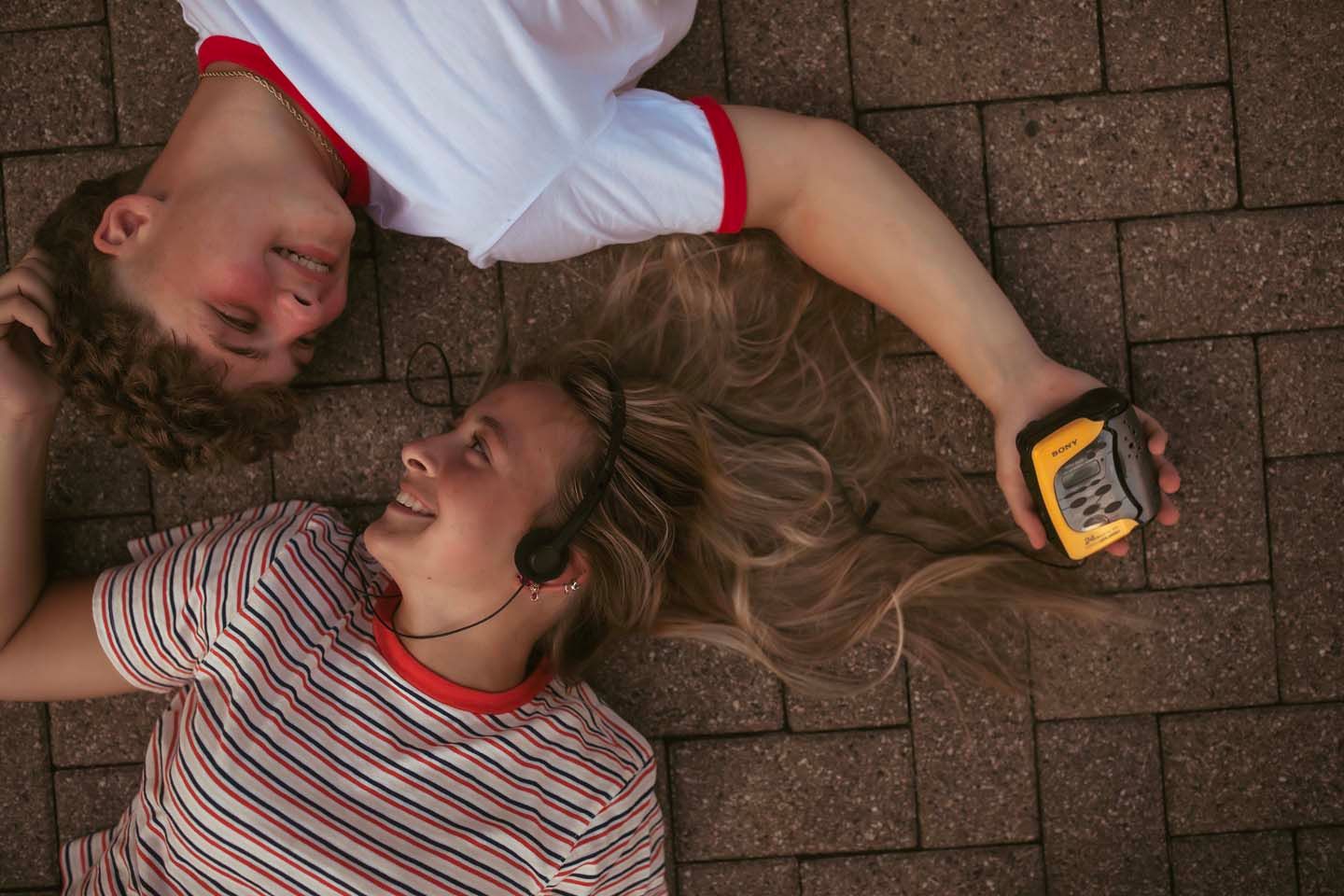 Overhead view of a young couple lying on brick pavement, sharing earphones and listening to a yellow music player.