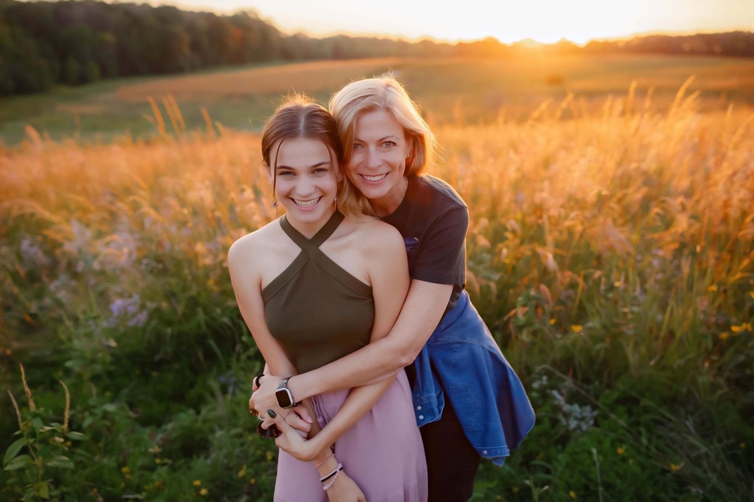 Two friends embrace and smile in a golden field at sunset with warm glowing sunlight behind them.