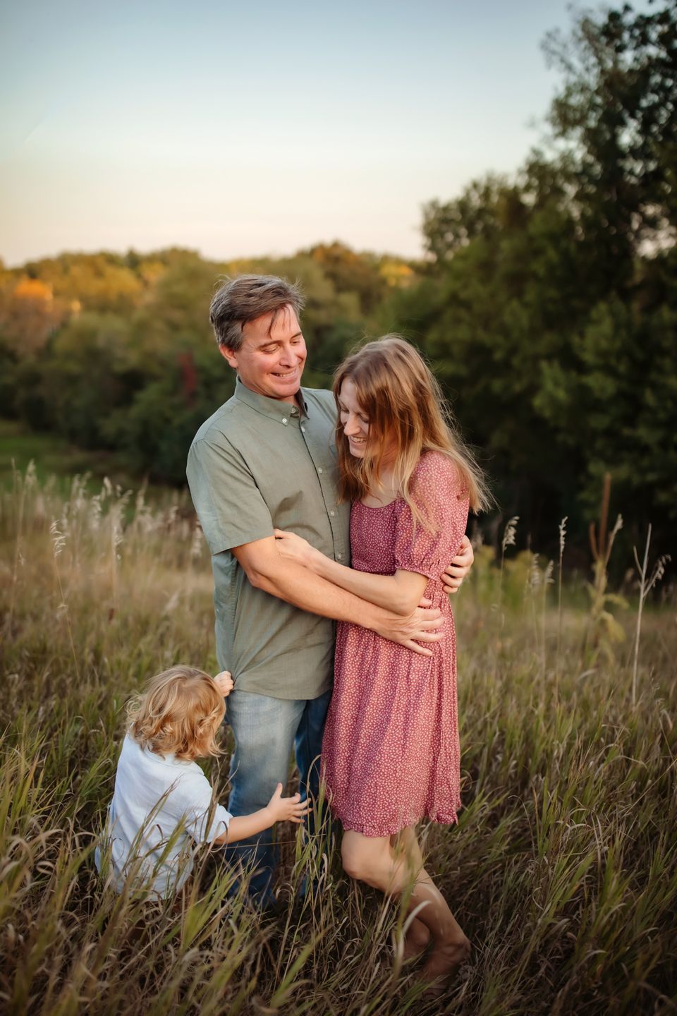 A loving family embraces together in a sunset meadow with tall grass and trees in the background.