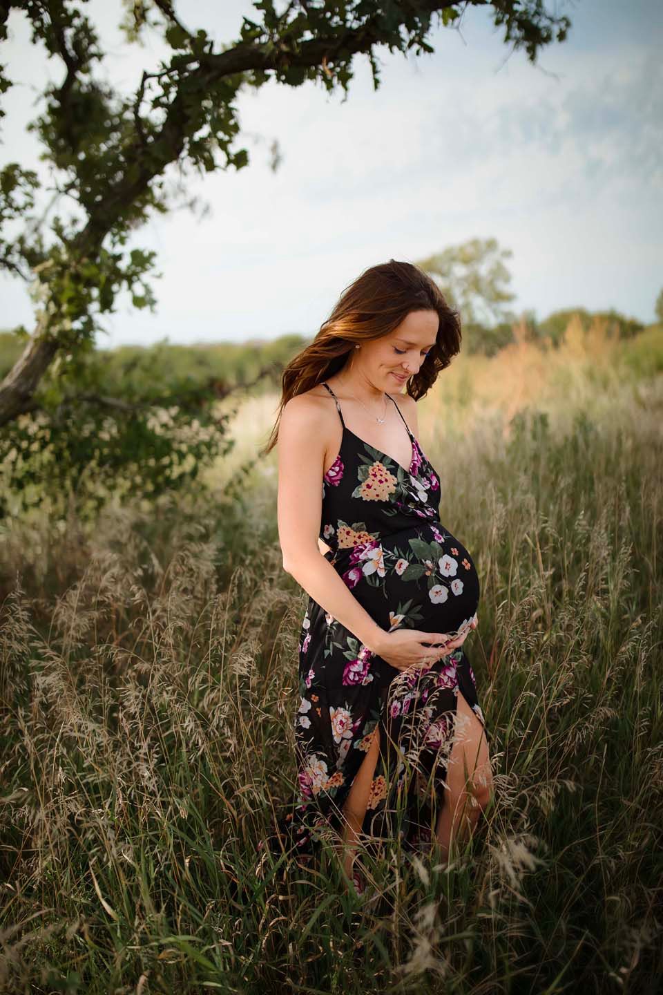 A pregnant woman in a floral dress stands in a sunlit meadow, smiling while holding her baby bump.