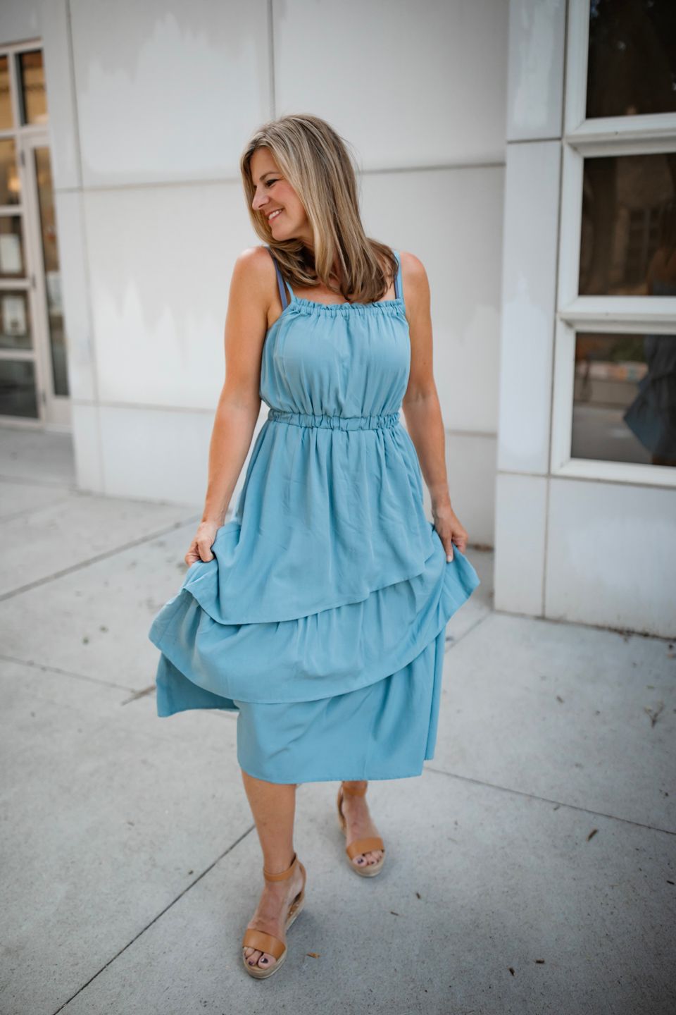 Casual fashion shot in a flowy blue summer dress walking along white exterior wall.