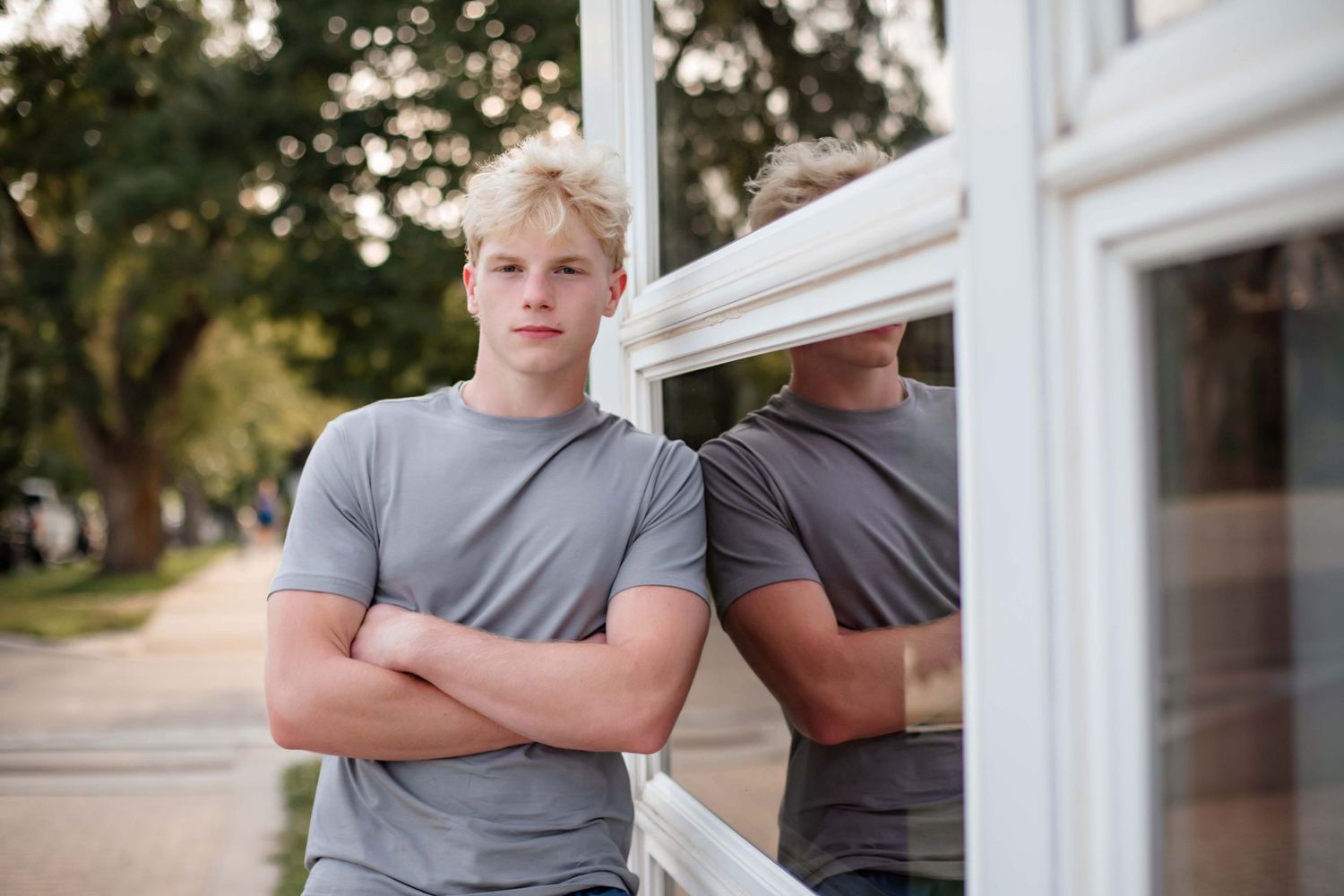 Casual pose against white window frames with reflection visible.