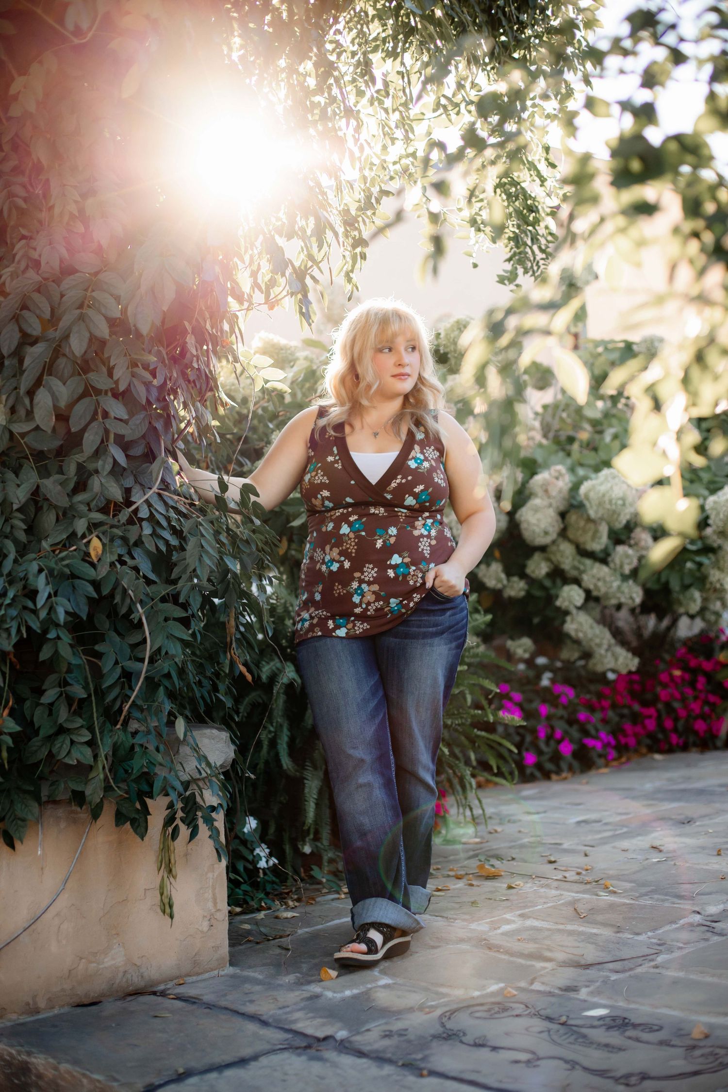 Outdoor portrait with sun flare and pink flowers beside stone pathway.