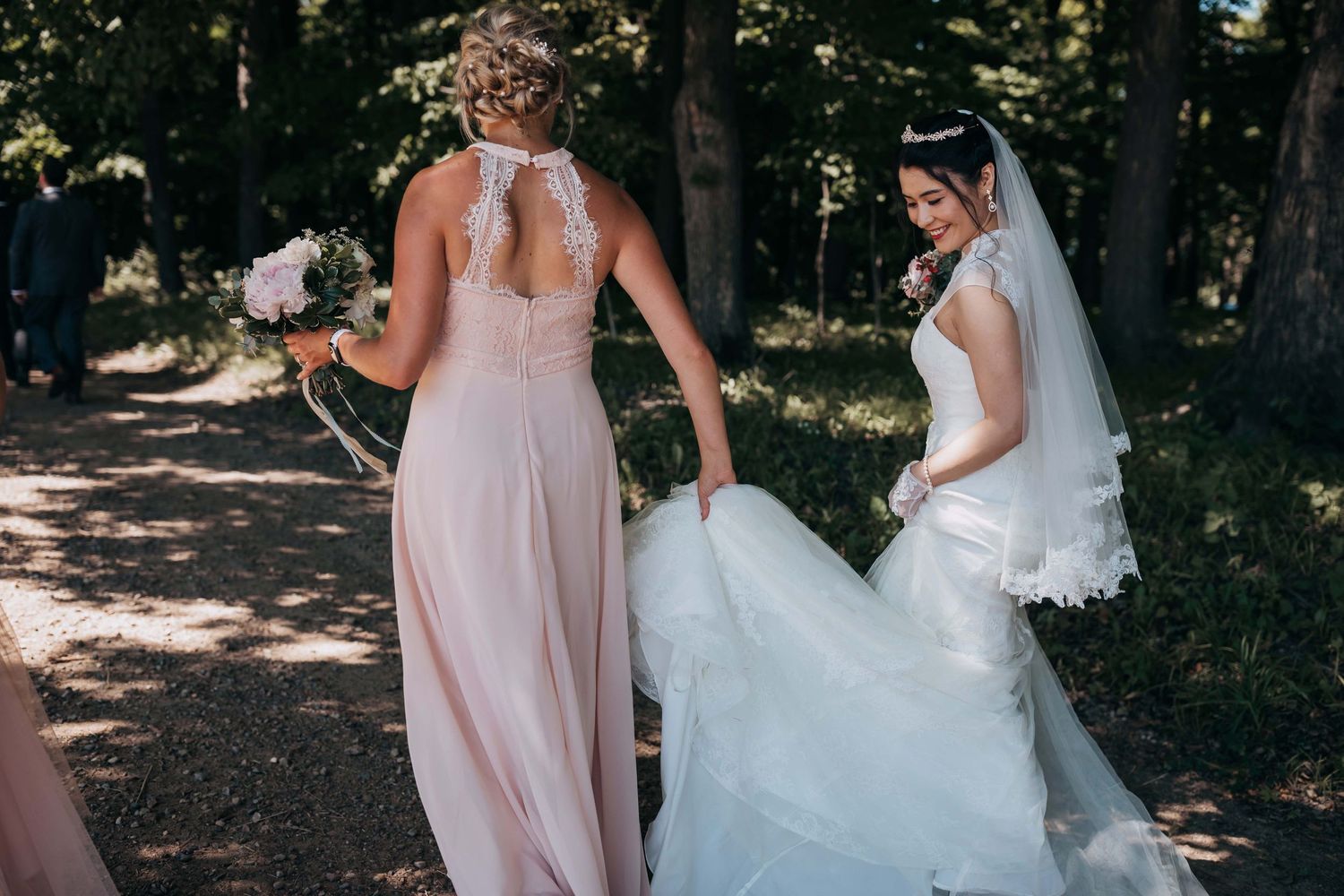 Bridesmaids in pink dresses help the bride with her flowing white wedding gown outdoors.