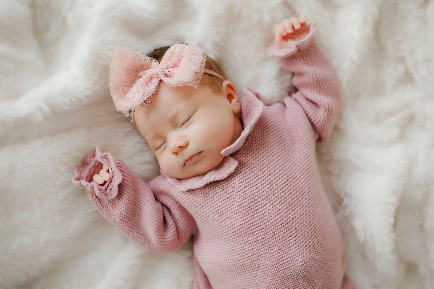 Baby wearing pink sweater and bow sleeping peacefully on white blanket.