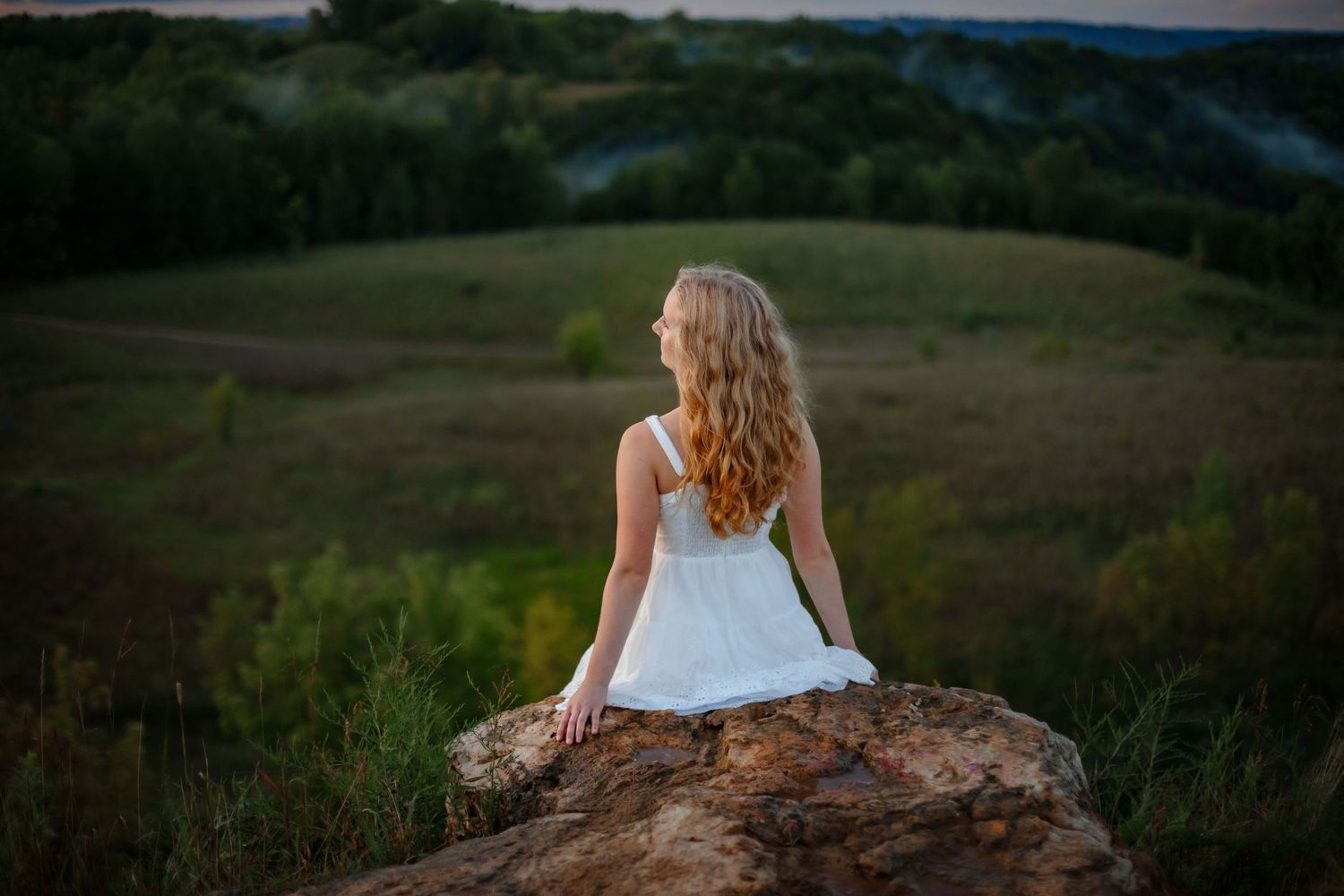 A white dress contrasts against a sunset landscape as someone sits perched on a rock overlooking rolling hills.