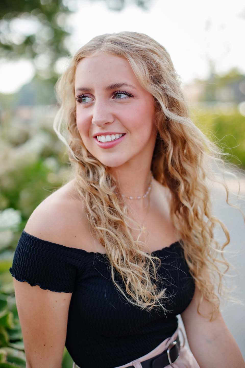Summer portrait with wavy blonde hair wearing a black off-shoulder top in a garden setting.