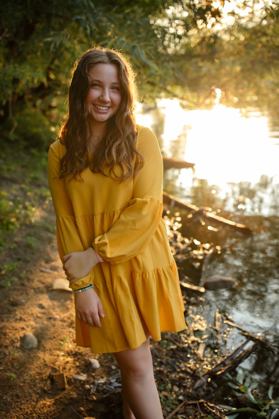 Person wearing a flowing yellow dress poses by a river at golden hour.