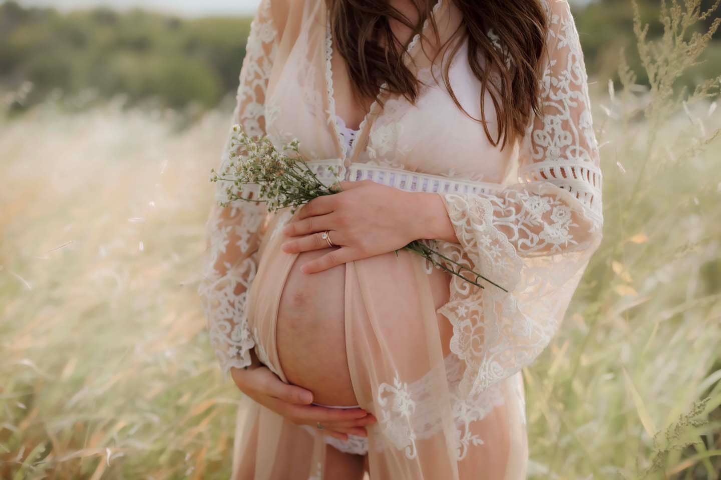 Expecting mother in lace dress cradles baby bump while holding wildflowers in golden sunlit meadow.