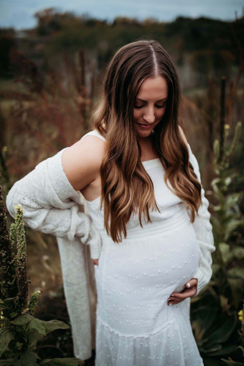 A pregnant woman in a white lace dress and cardigan stands peacefully in a garden setting at sunset.