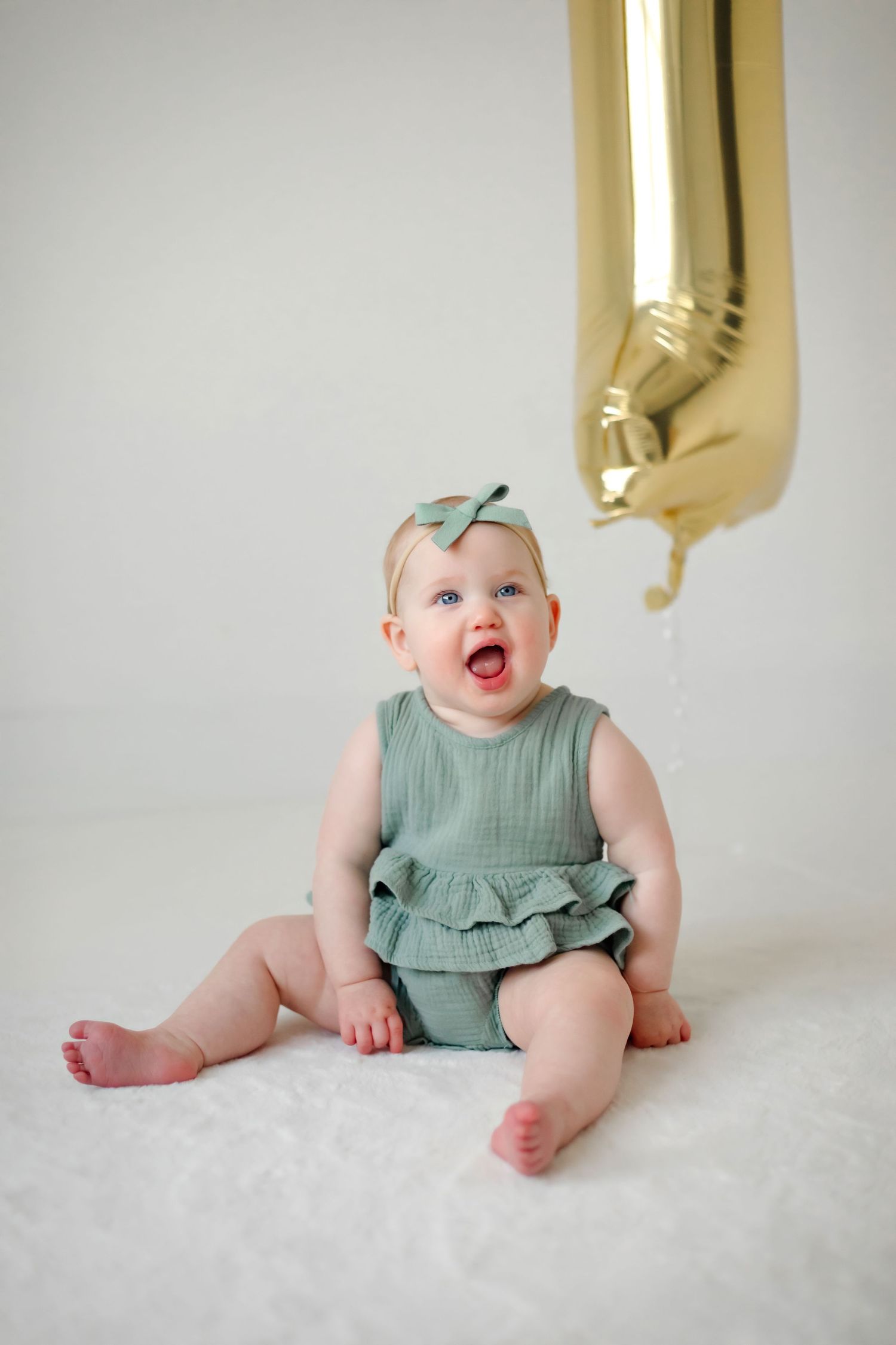 Baby in mint green ruffled romper sitting beneath gold balloon on white backdrop.