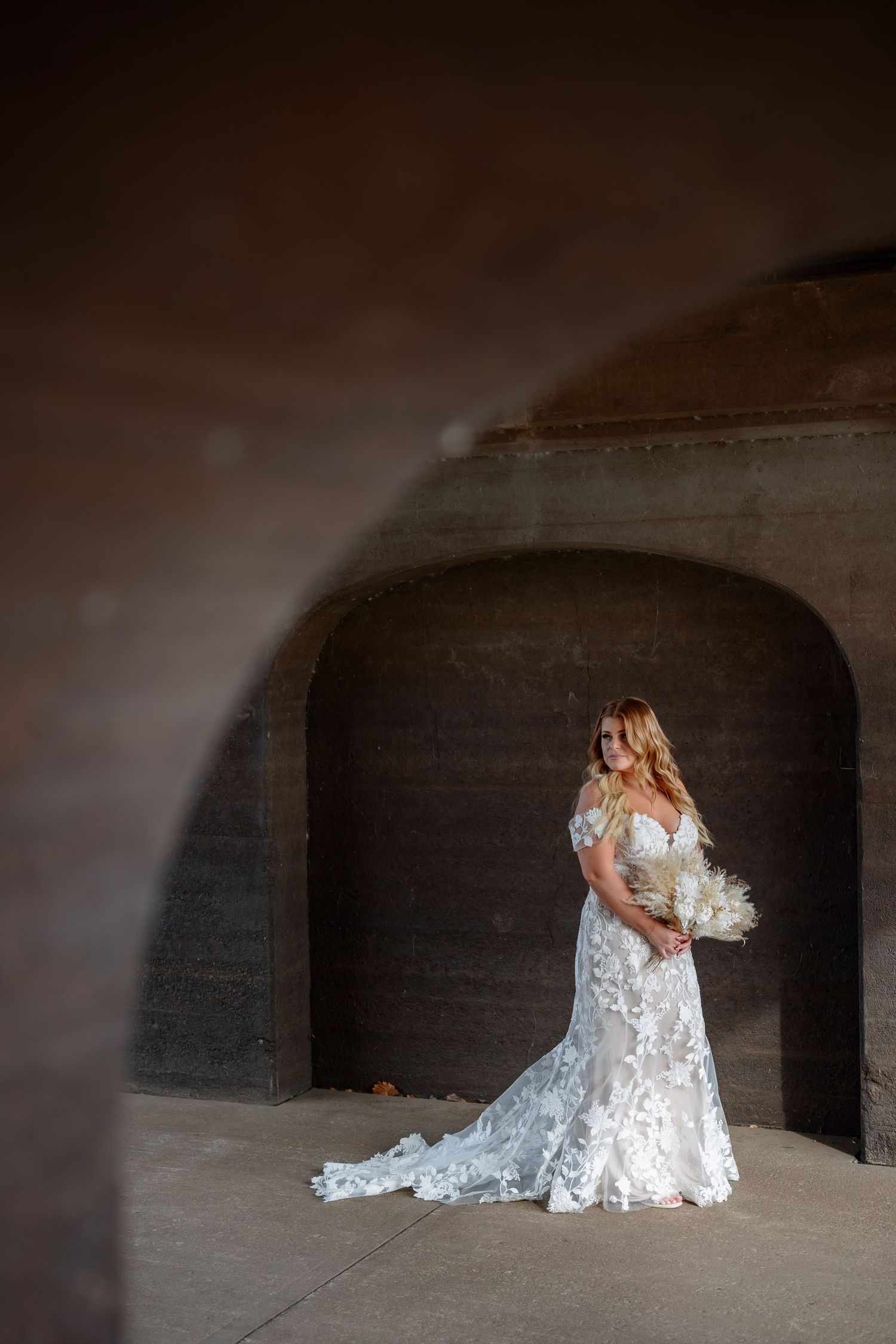 A bride in a lace wedding gown poses in a dramatic stone archway with natural lighting.
