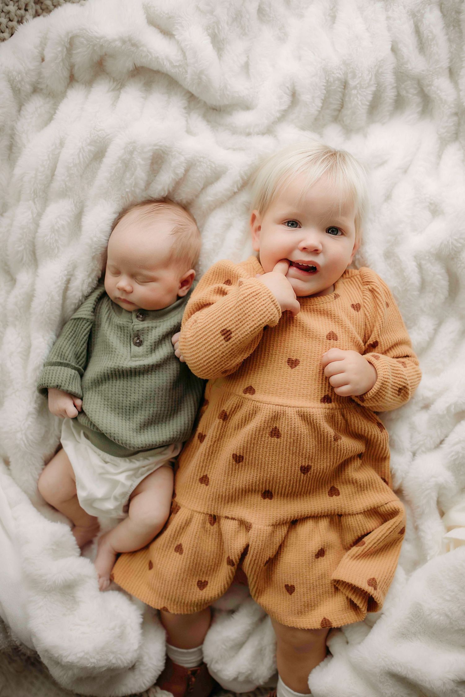 Two babies lying on plush blanket wearing knit outfits in green and mustard colors.