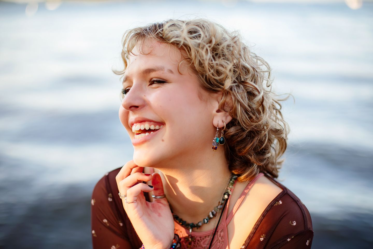 A person with curly blonde hair laughs joyfully outdoors at sunset wearing a burgundy top and turquoise earrings.