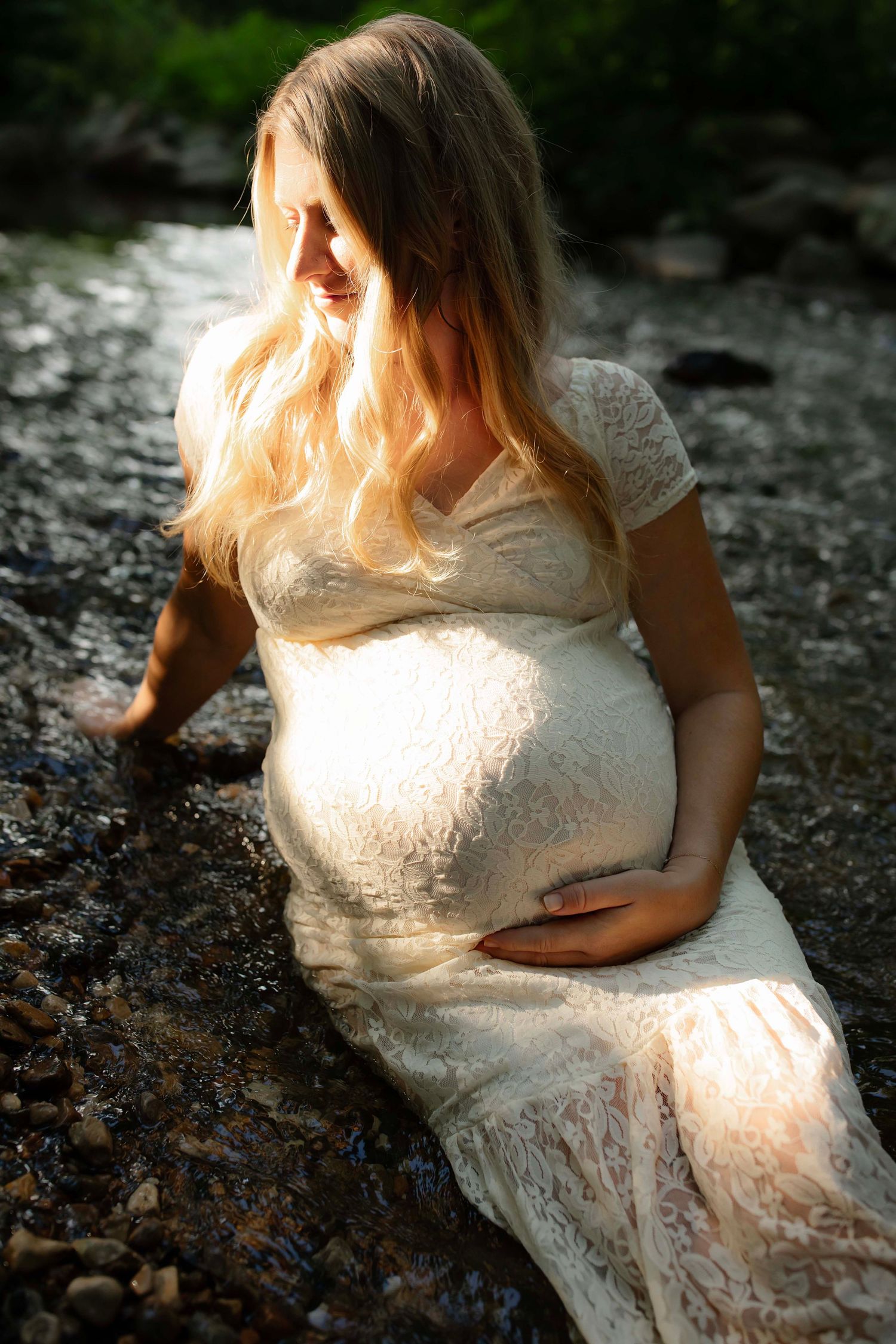 Pregnant woman in white lace dress sitting by a rocky stream, cradling her baby bump in golden sunlight.