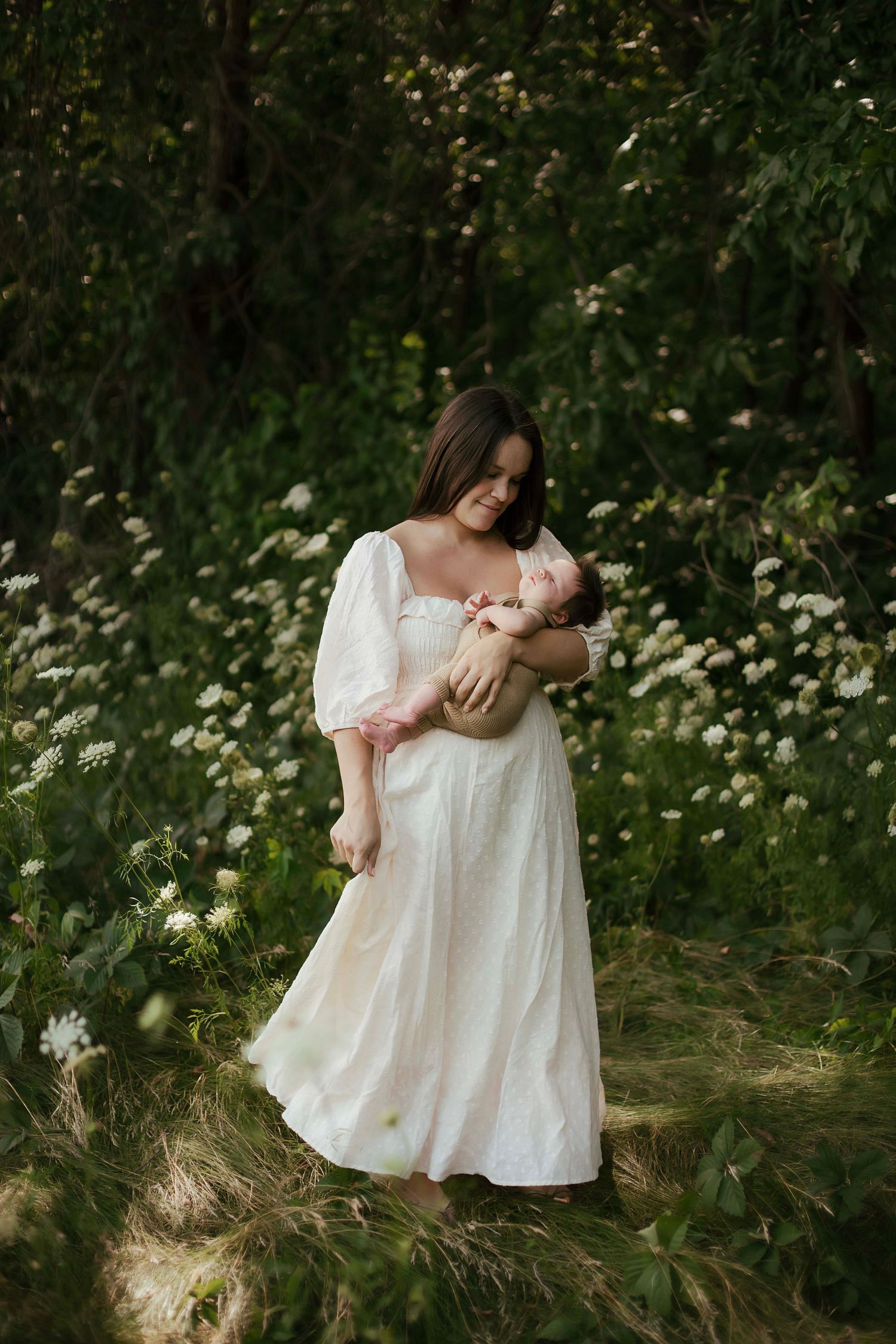 Mother in white dress cradling newborn baby outdoors among wildflowers and lush green trees.