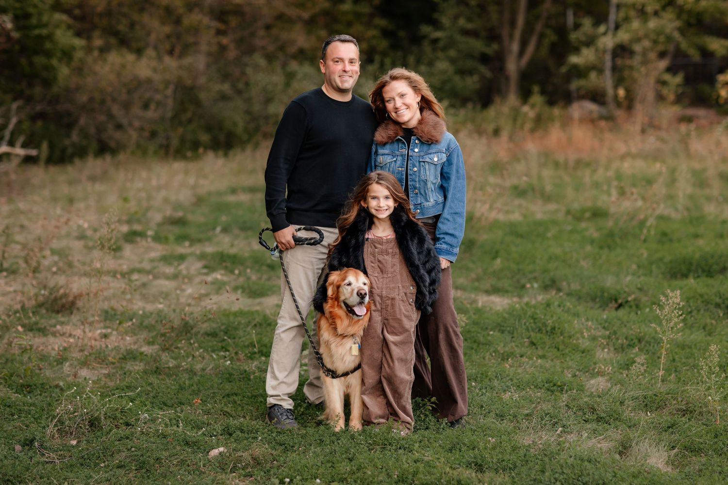 Family of three with golden retriever dog posing outdoors in a green field surrounded by trees.