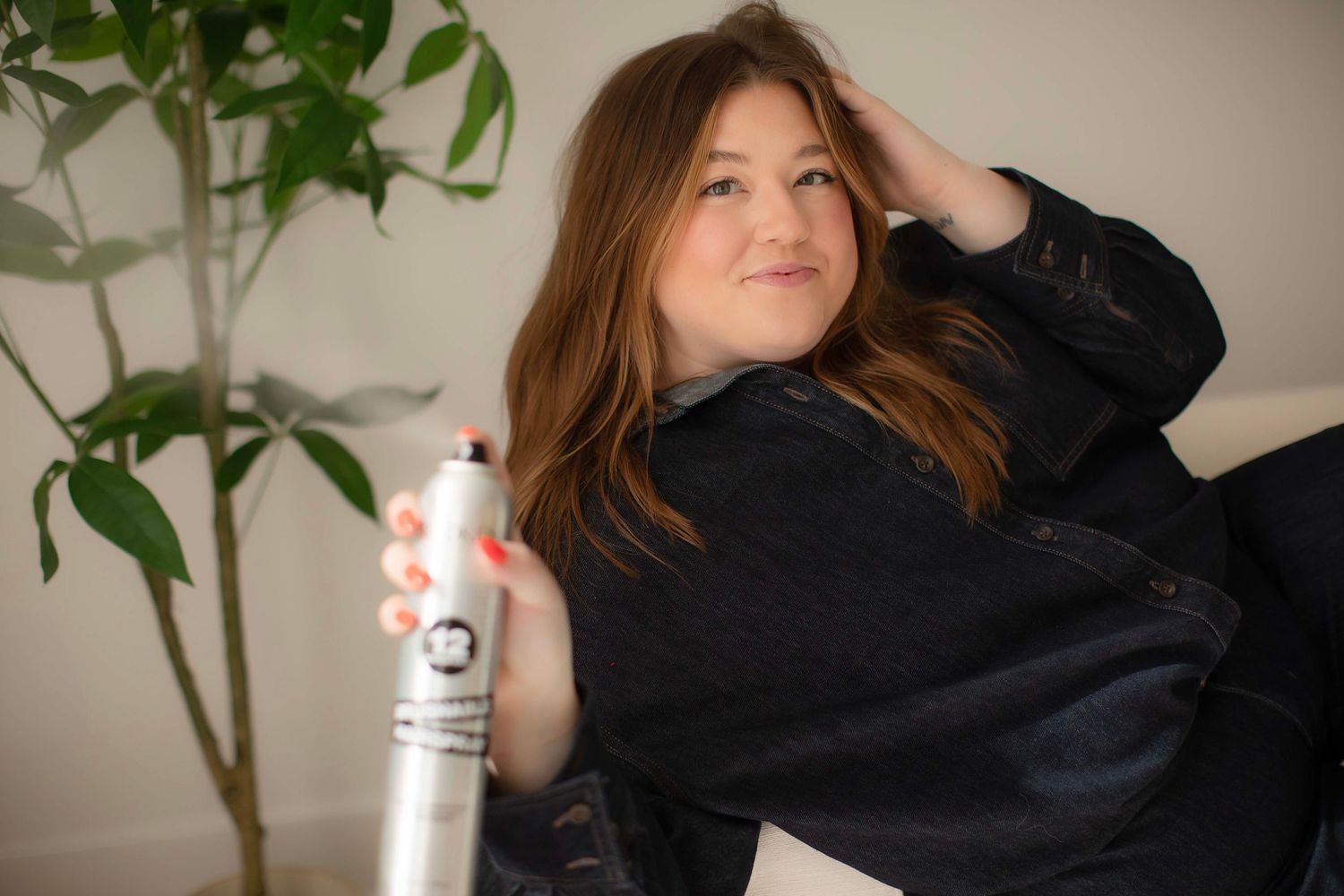Woman holding a hair spray can while touching her hair, wearing a black jacket, seated near a green plant.