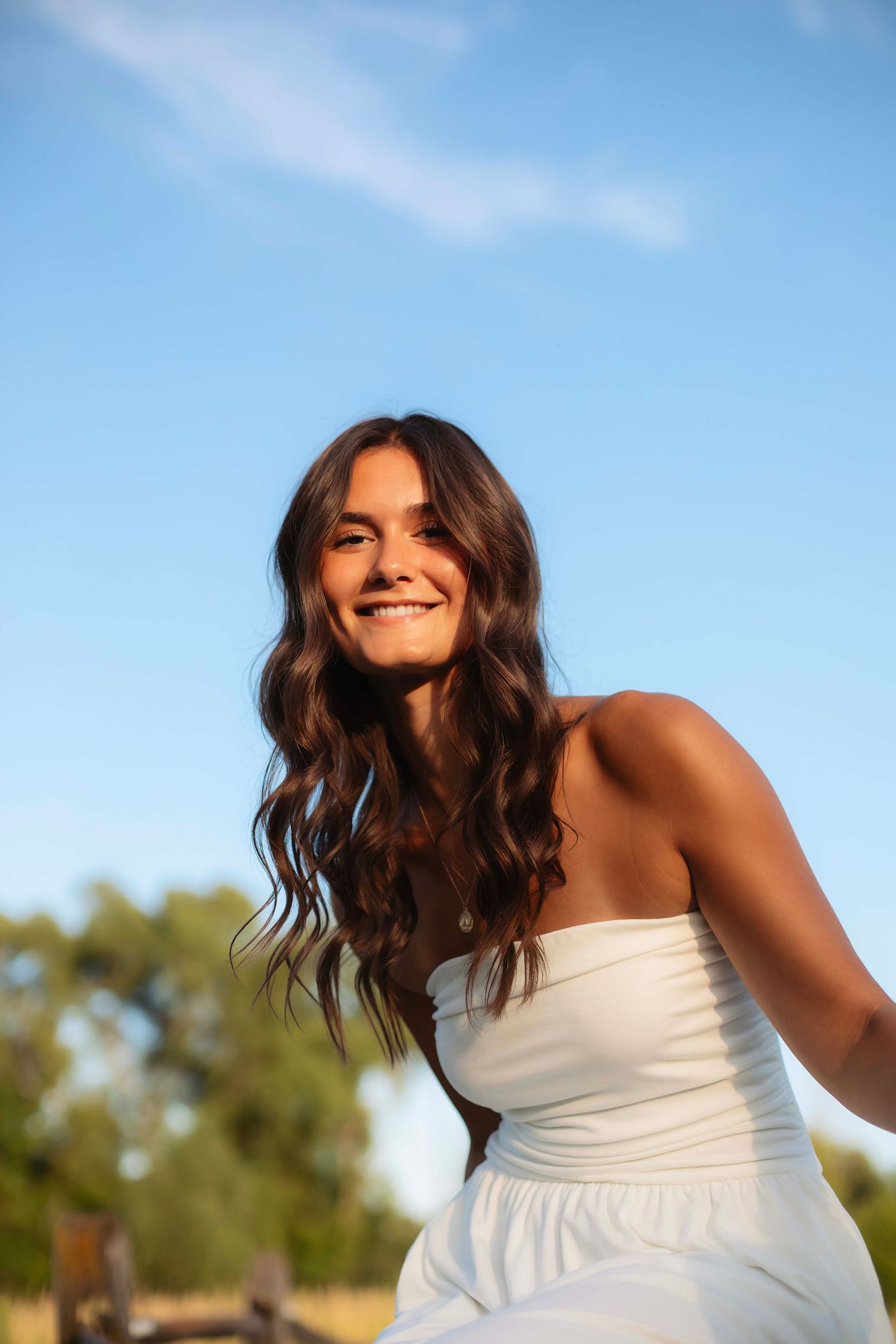 Young woman with long wavy brown hair smiling outdoors in a white strapless dress against a blue sky.