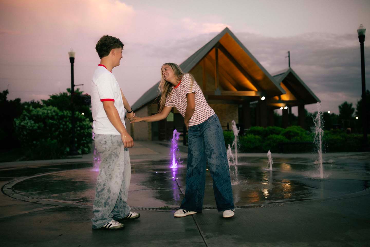 A young couple plays near illuminated fountains at dusk outside a modern building.