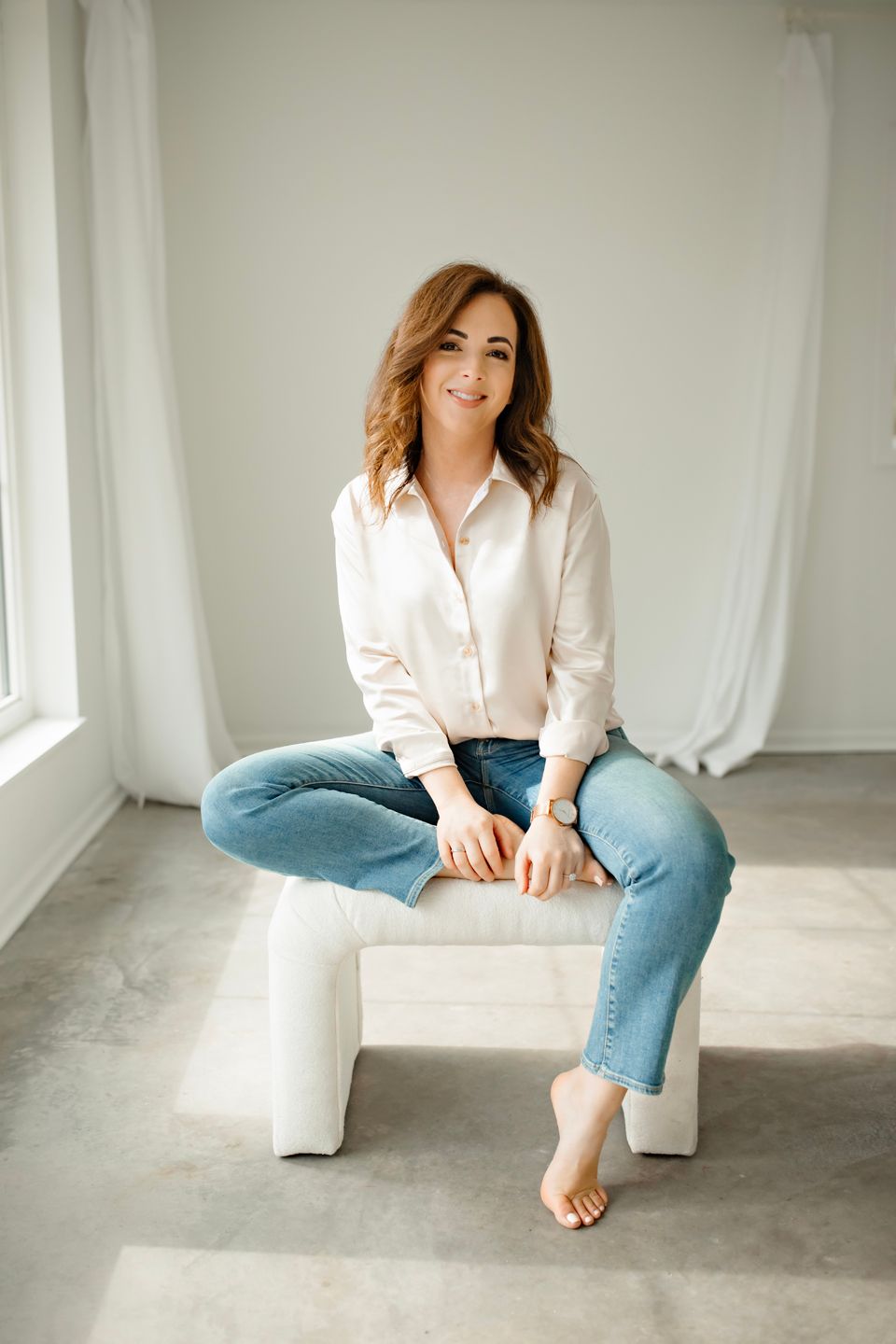 A woman in a white button-down shirt and blue jeans sits casually on a white bench against a light backdrop.
