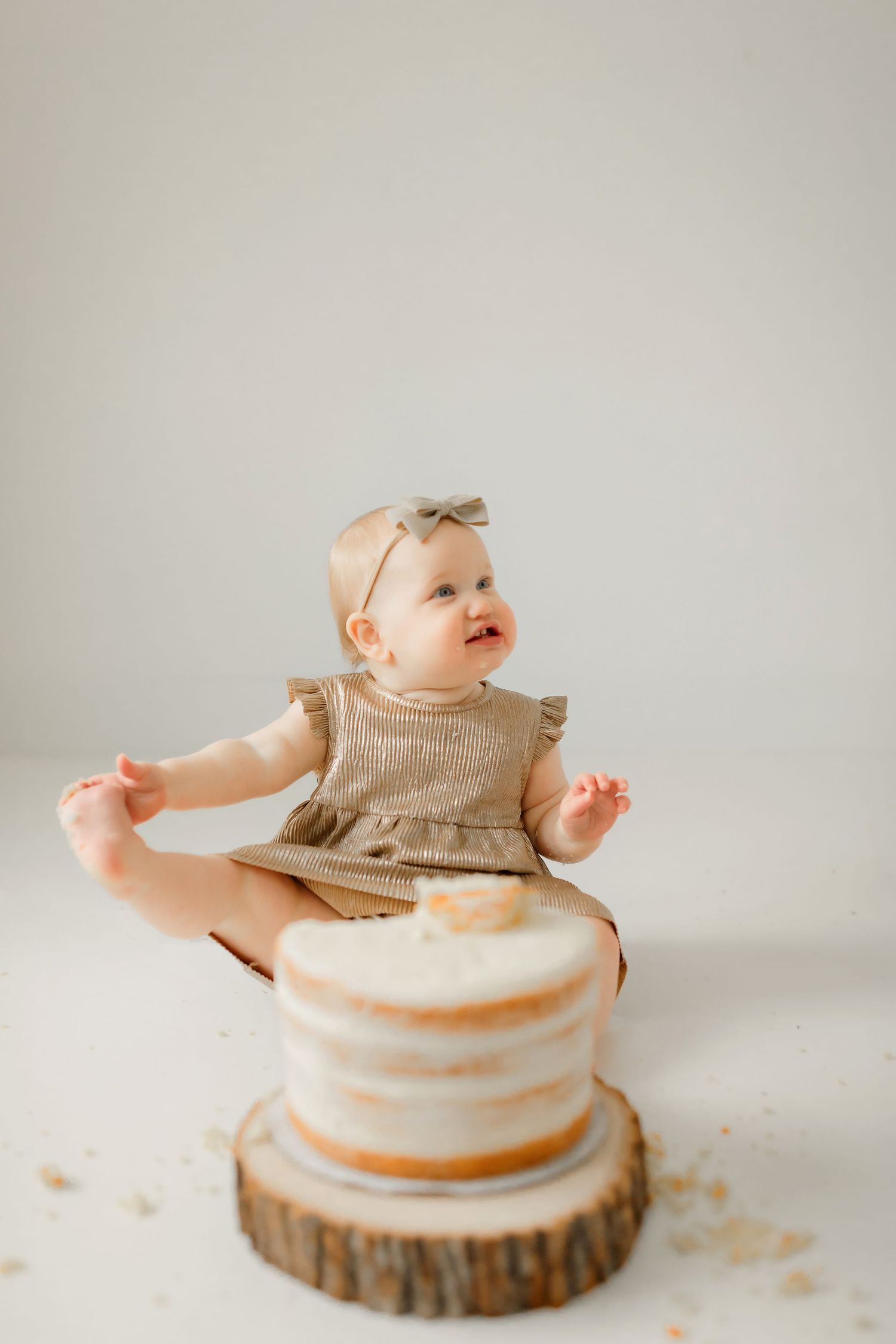 A baby in a beige dress sits next to a rustic tiered cake on a wooden slice against a neutral background.