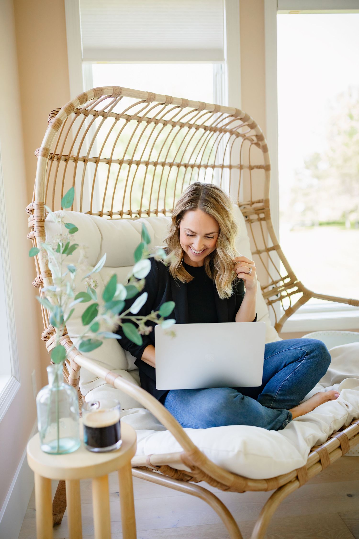 Person working on laptop in comfortable rattan hanging chair with decorative plants and side table.