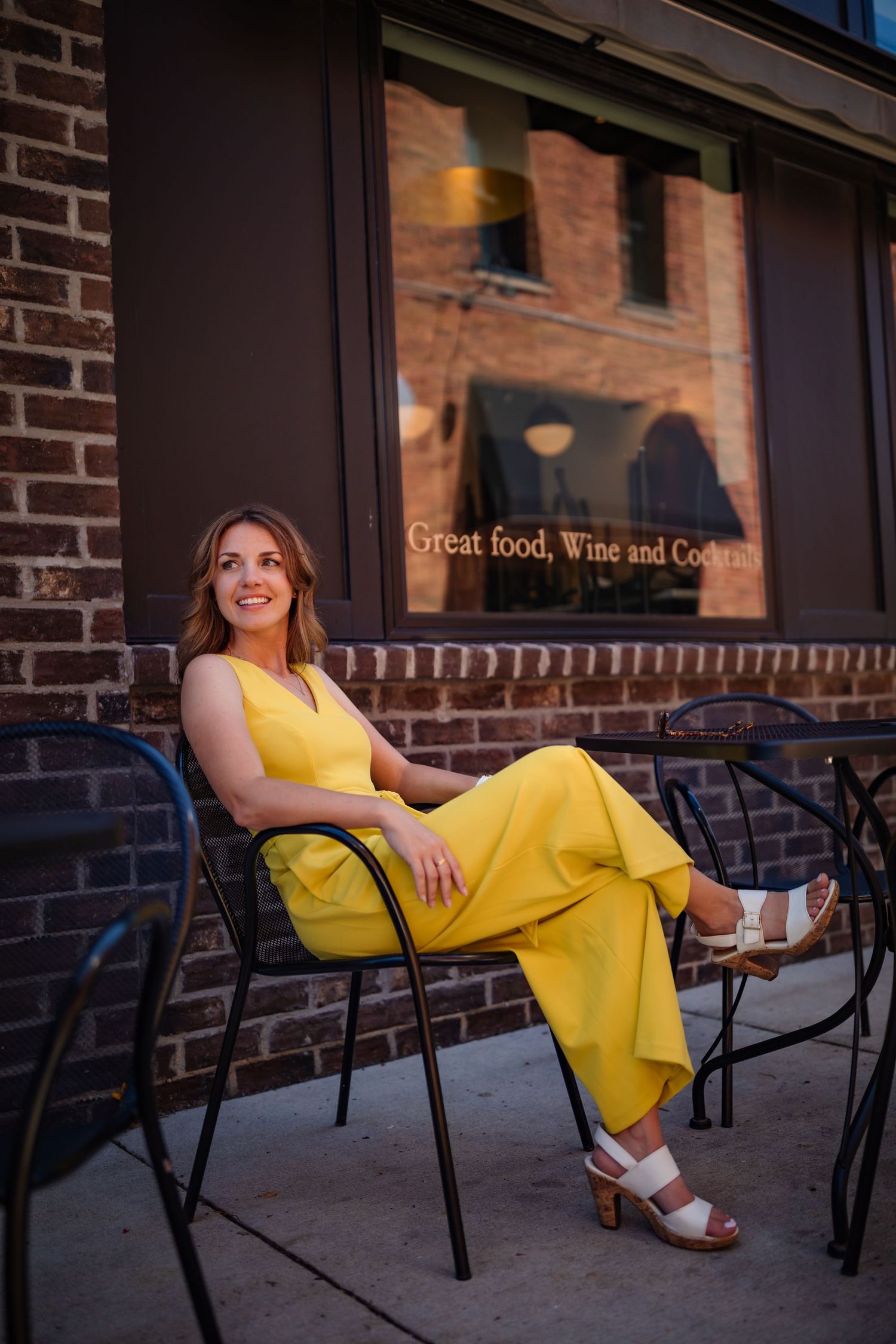 Woman in yellow jumpsuit sits relaxed at outdoor cafe table near brick building.