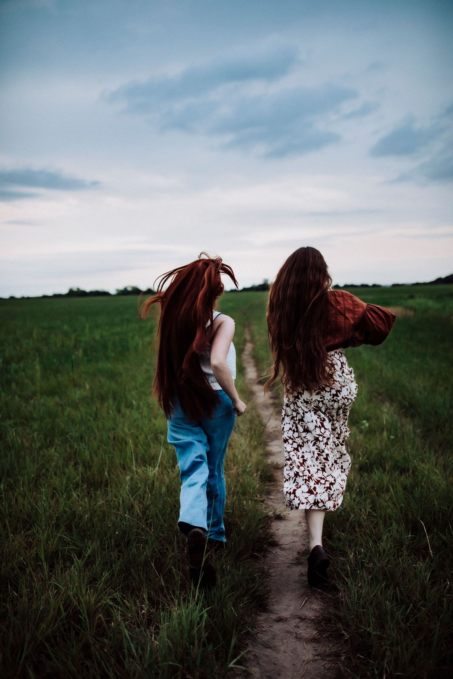 Two people with long red hair walking together down a dirt path through a green field.