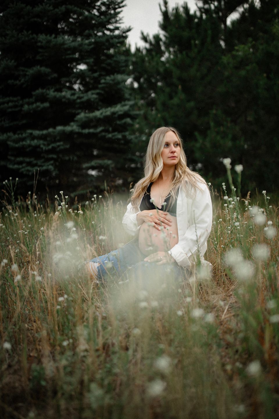 A maternity photo taken in a dreamy wildflower field with evergreen trees in the background.