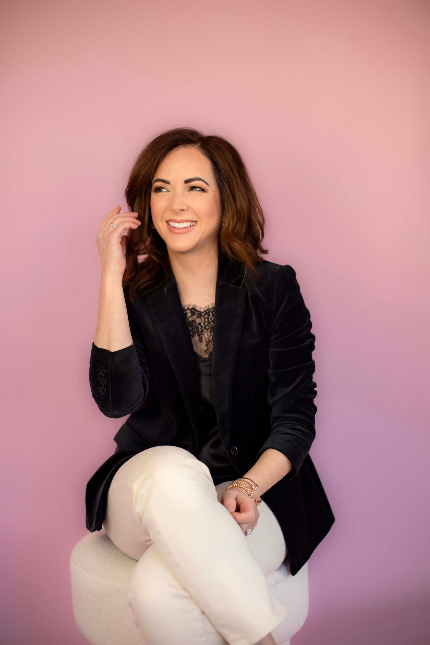 Seated casual business portrait with black blazer and lace top against pink gradient background.