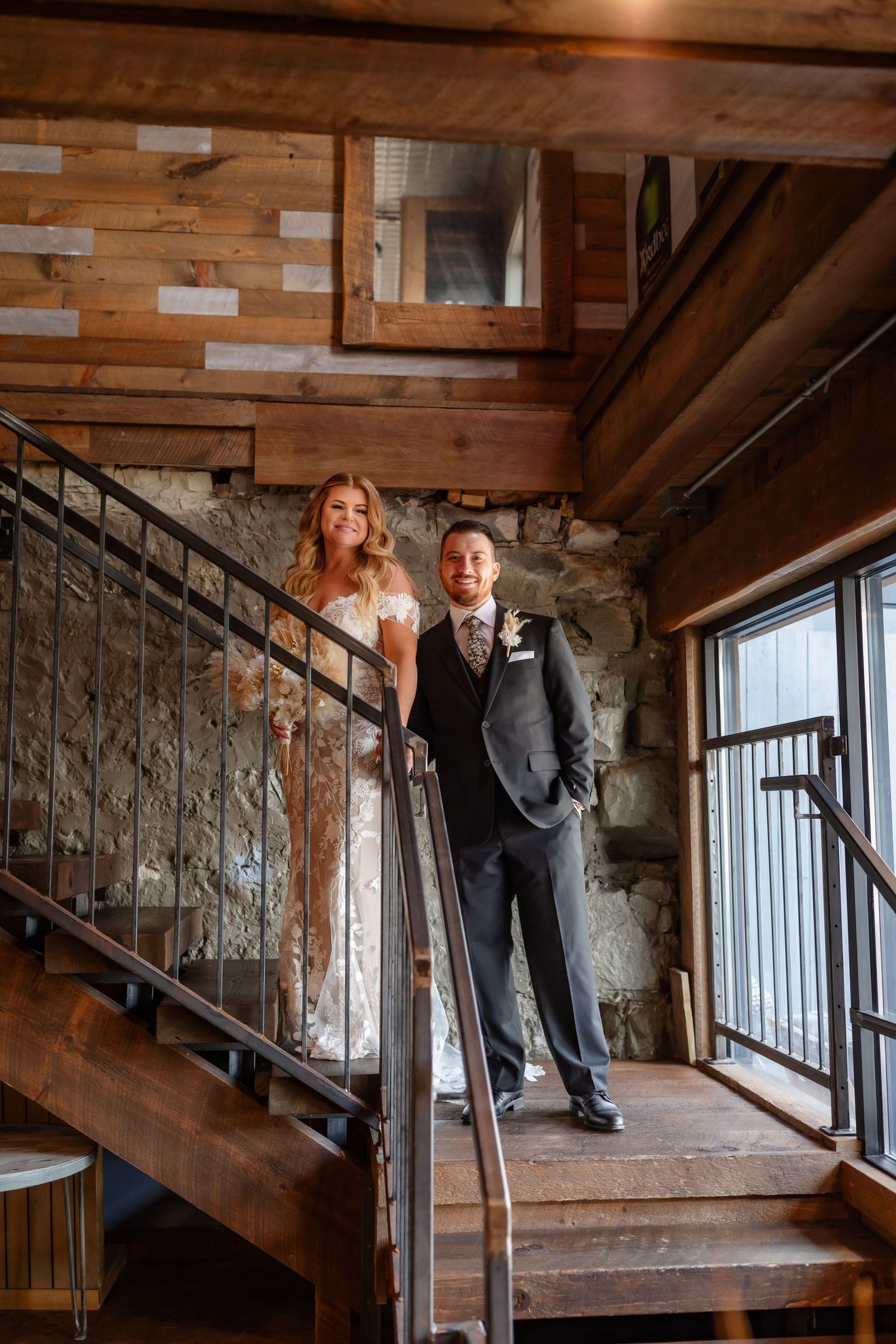 A couple poses on wooden stairs in a rustic venue with stone walls and wooden beams.