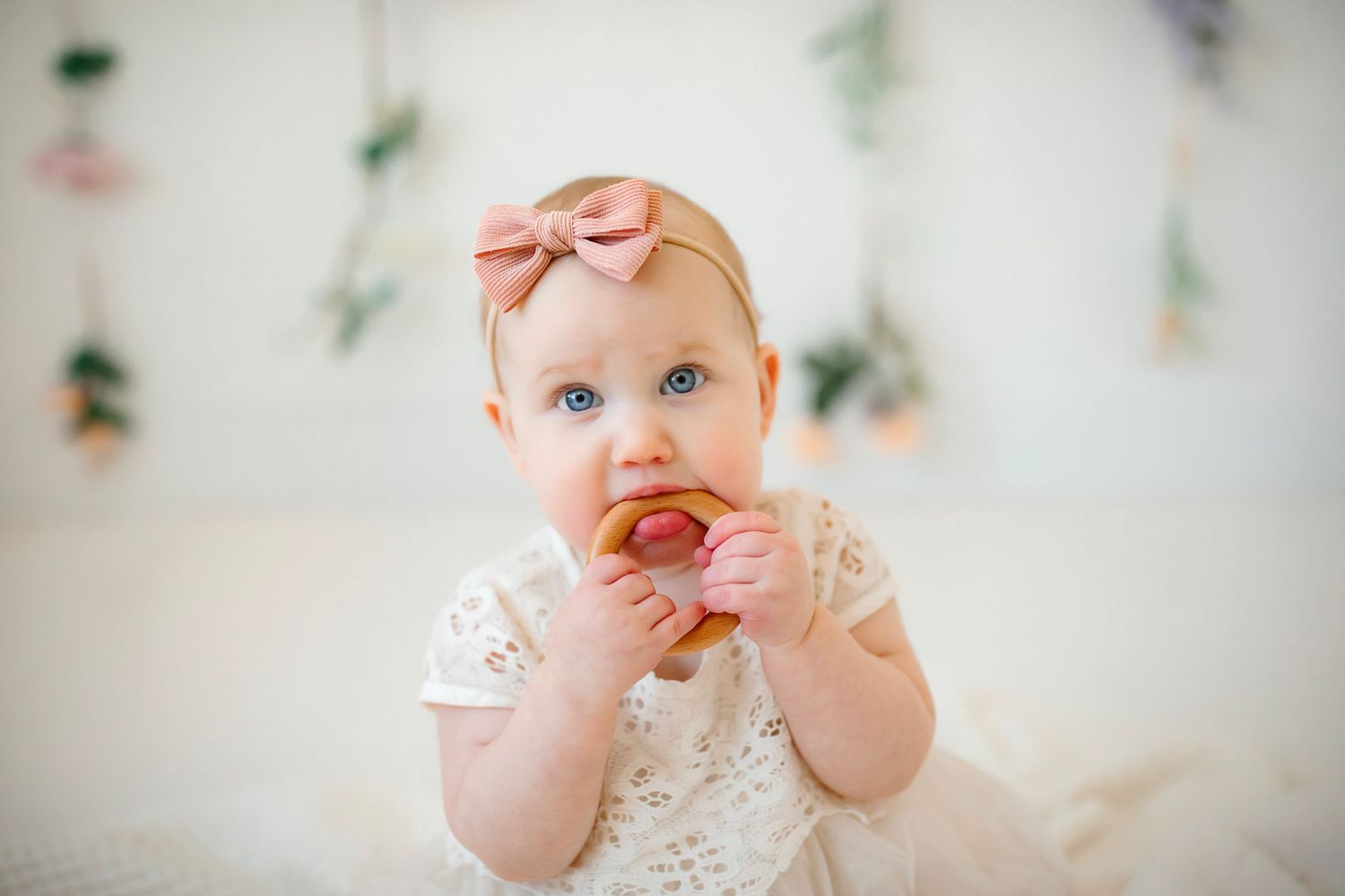Baby wearing white lace dress and peach bow headband sitting against floral backdrop.