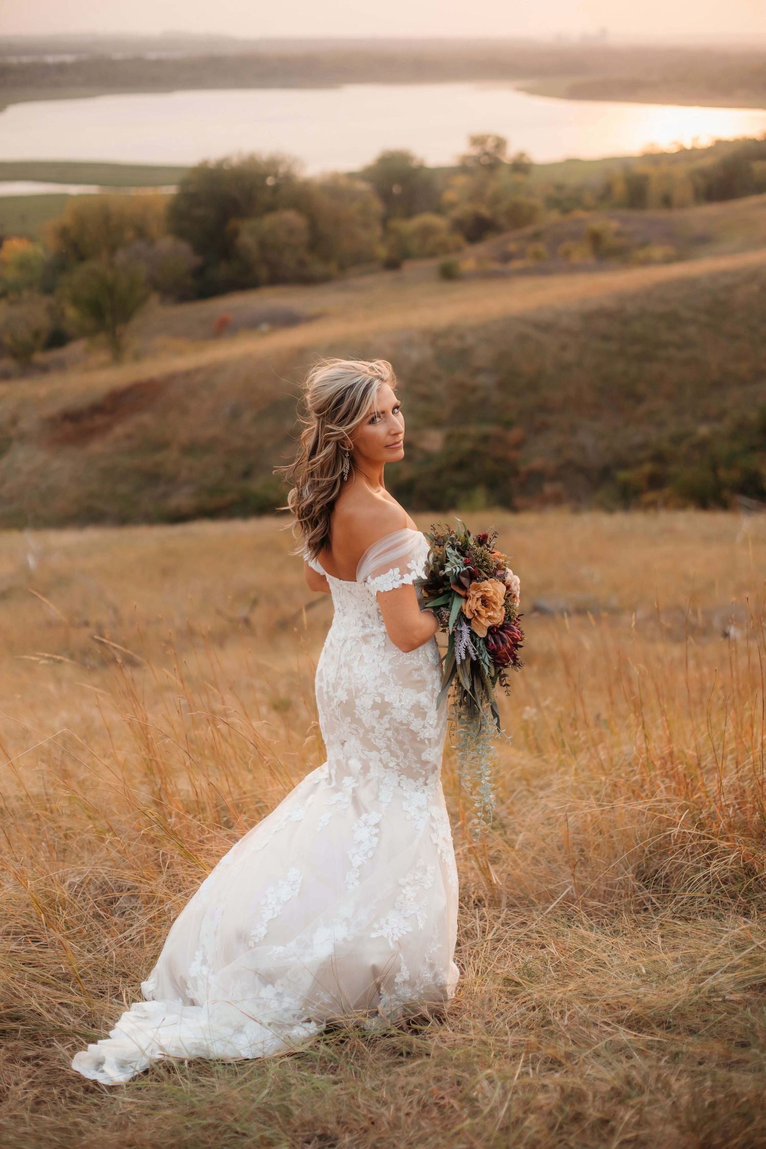 A bride in white lace stands against a scenic lake view during golden hour.