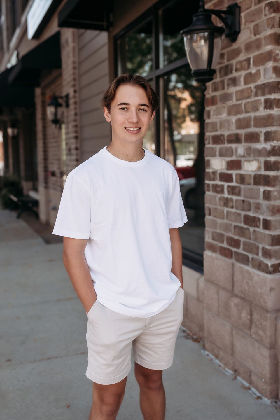 Person in casual white t-shirt and khaki shorts smiling while standing against brick wall on downtown sidewalk.