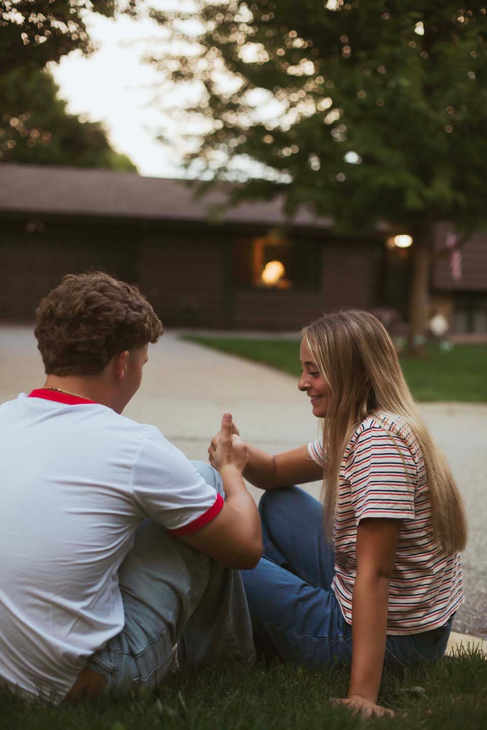 A young couple sits on grass at dusk, smiling and holding hands near a cozy house with trees in the background.