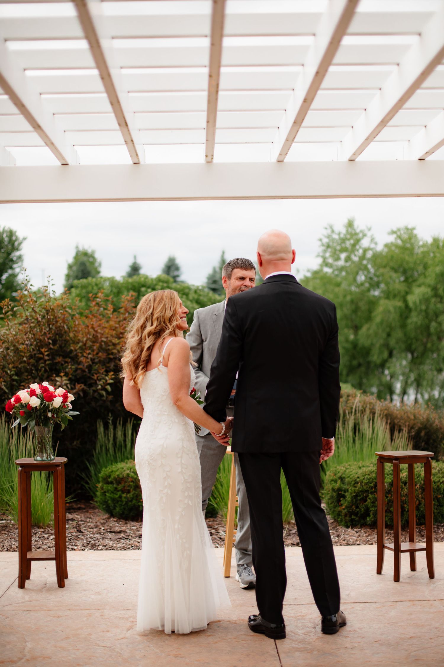 A wedding couple shares intimate moments under a white pergola with wooden stools and floral arrangements.