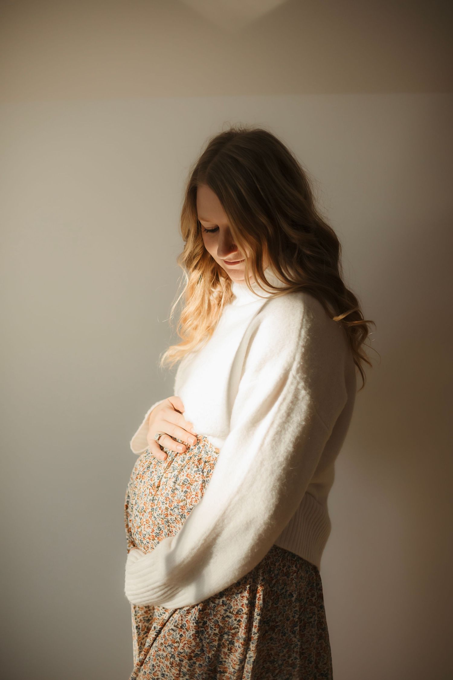 Pregnant woman in white sweater and floral skirt tenderly cradles her baby bump in soft natural light. Willow & Wren Photography maternity p