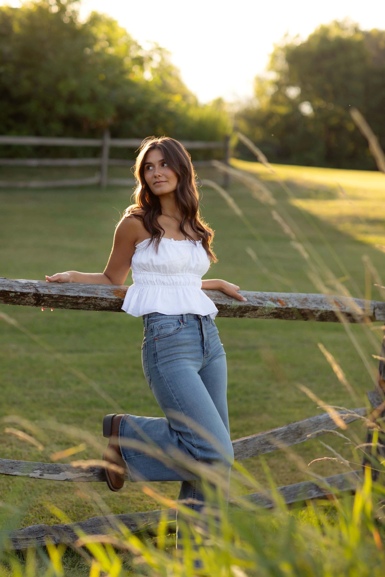 Young woman in white ruffled top and jeans leaning on wooden fence in sunlit green field.