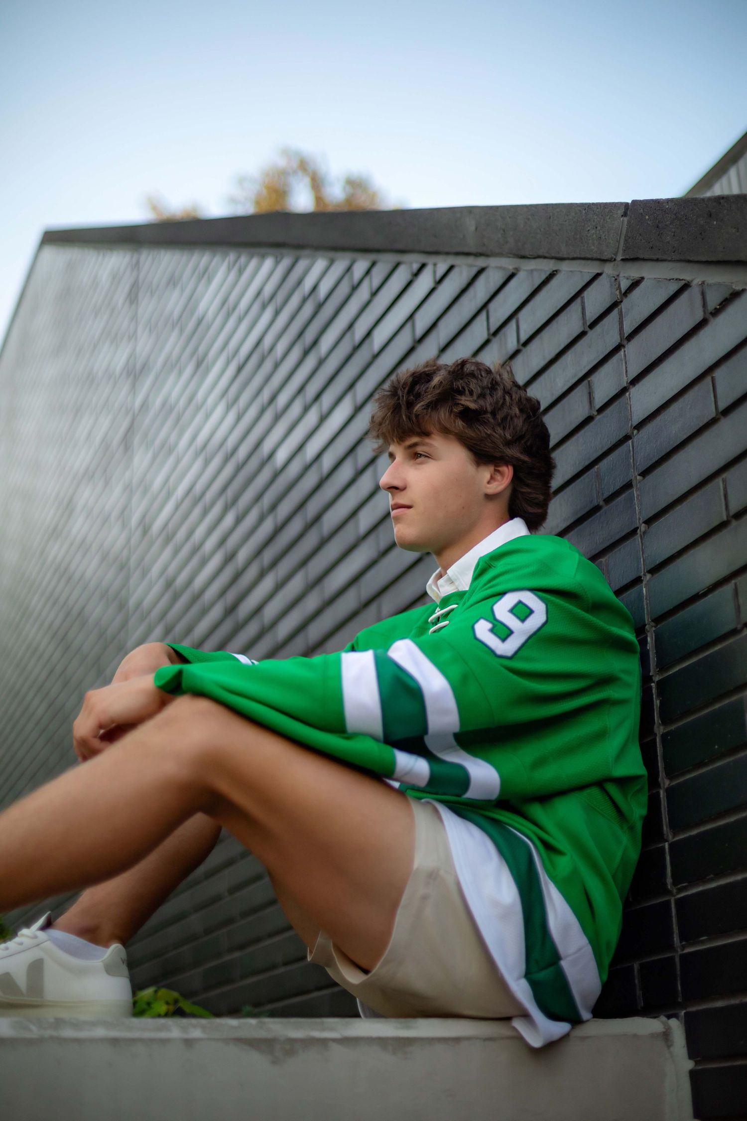 Young man in green jersey number 9 sitting outdoors against a dark metal wall, looking thoughtfully to the side.