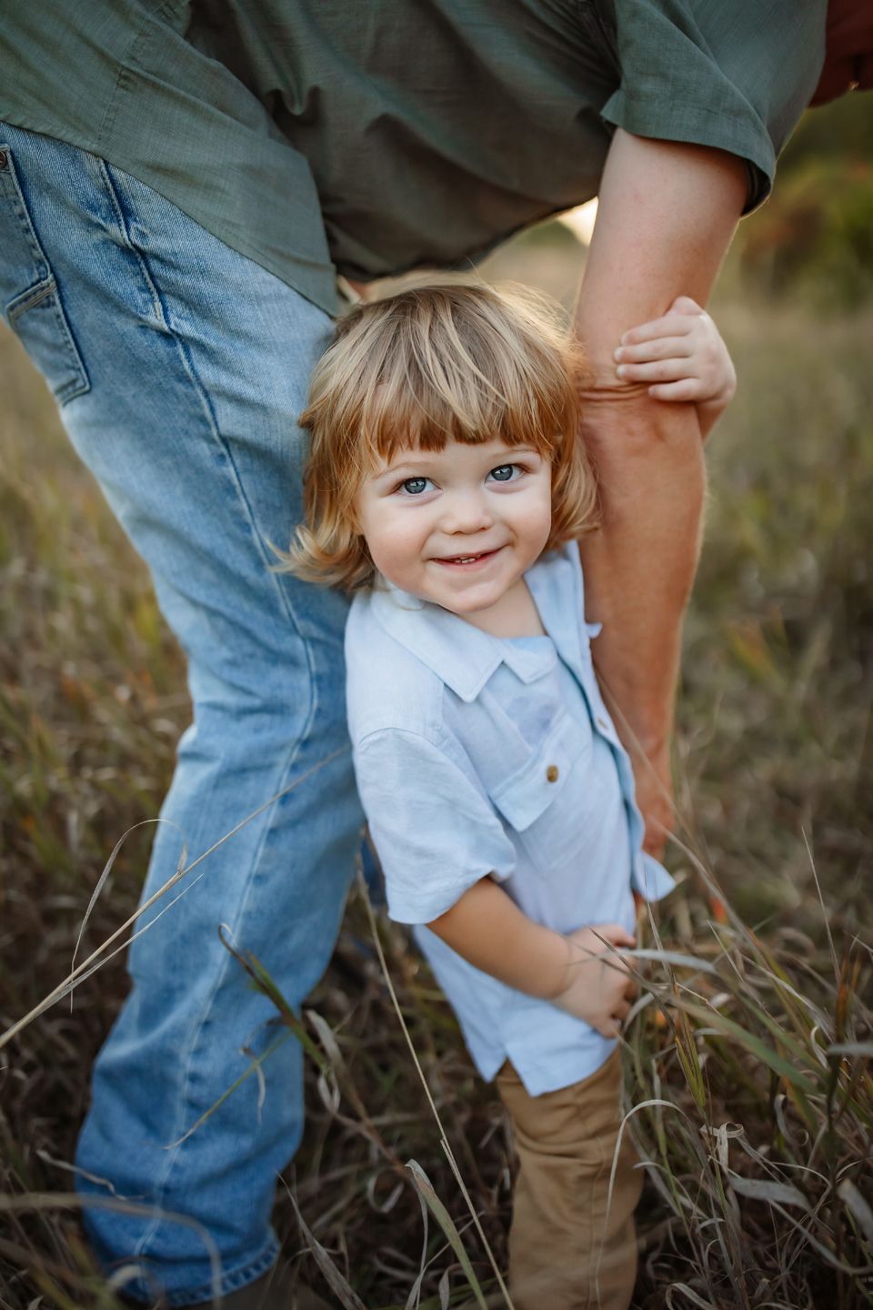 Small child in light blue shirt peeking out from behind adult's legs in a field.