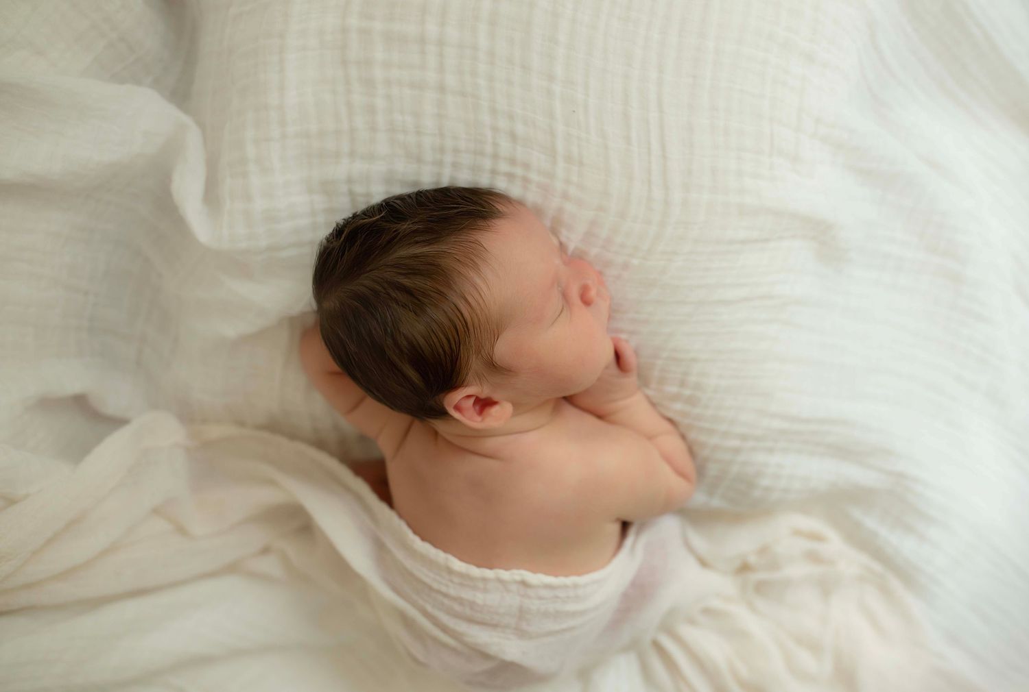 Peaceful newborn baby sleeping wrapped in soft white muslin cloth on a white fluffy blanket.