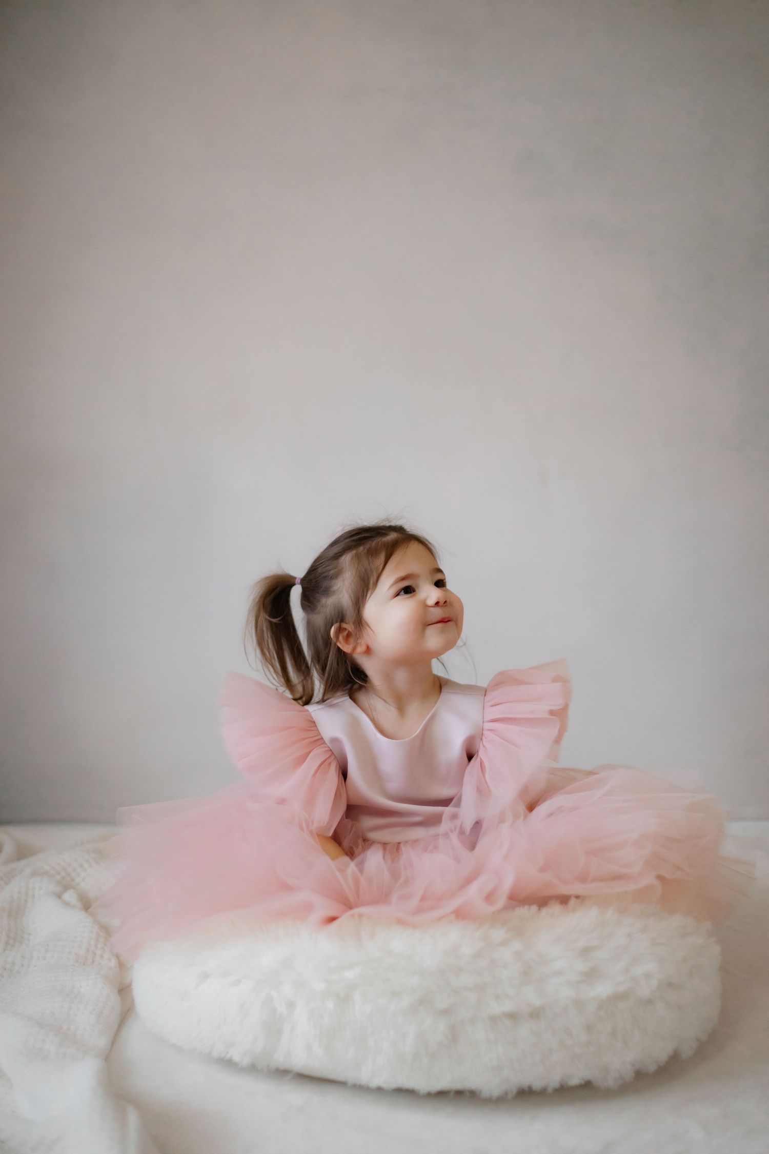 Child in pink dress sits on white furry cushion looking up dreamily.
