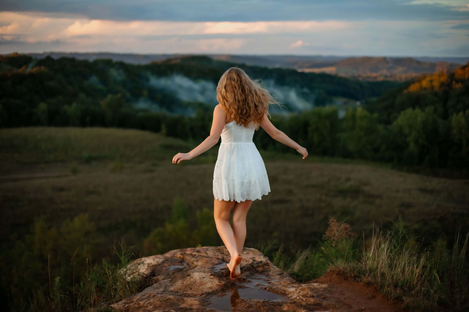 A carefree moment in a white dress on a clifftop overlooking rolling hills at sunset.