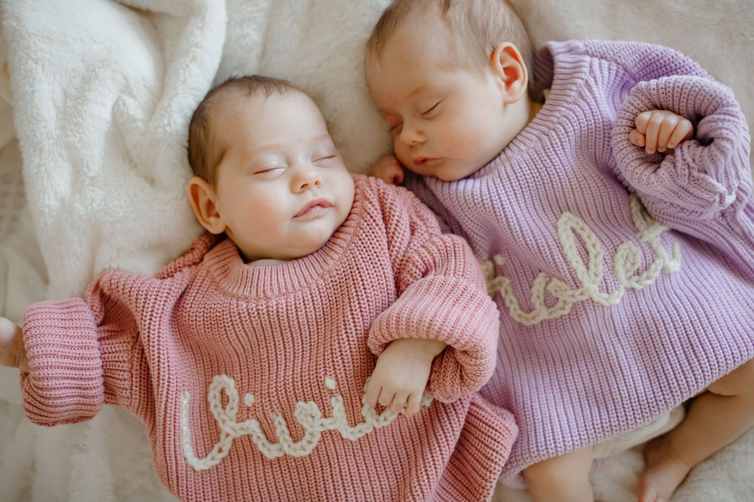 Twin babies sleeping side by side in matching pink and purple knit sweaters on white blanket.