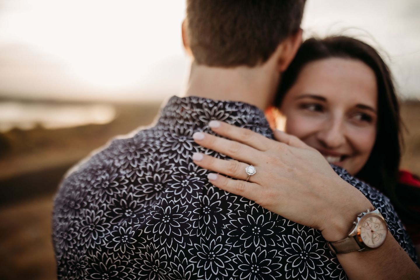 Close-up shot showing an embrace and engagement ring with a sunset landscape in the background.