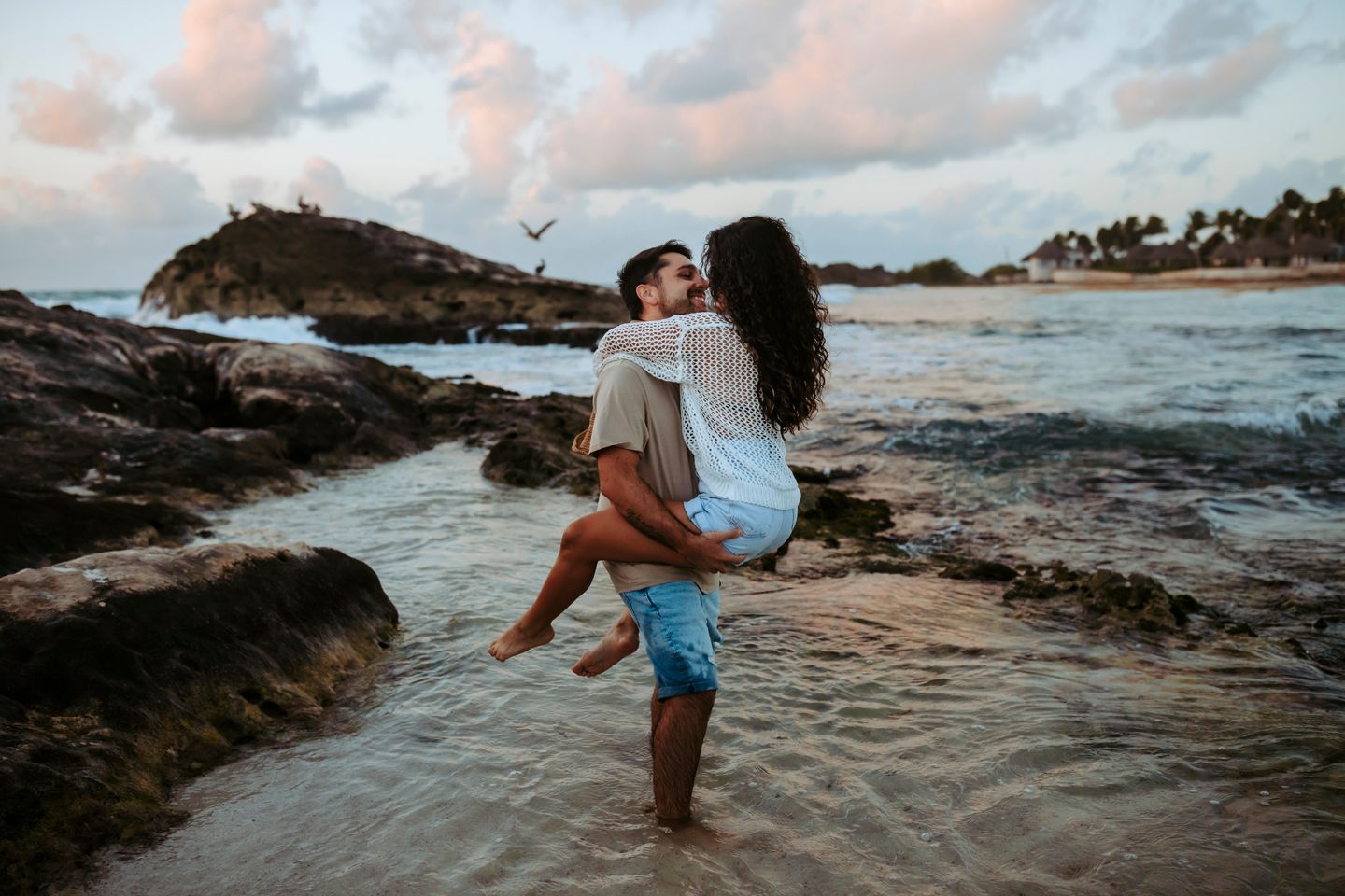 A couple sharing a playful moment in shallow water among coastal rocks at sunset.