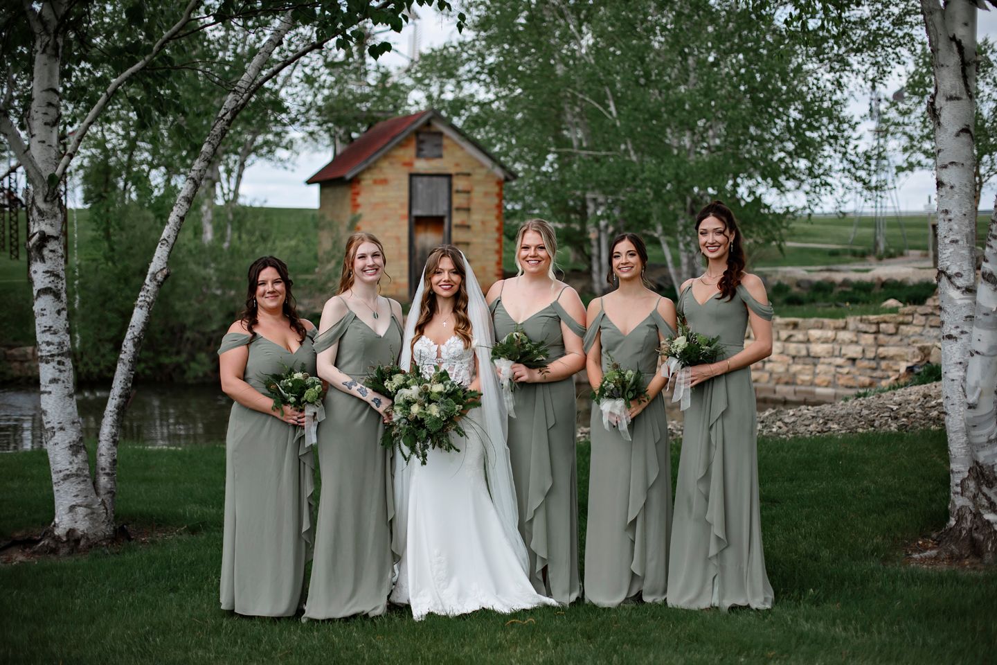 Bridal party in sage green dresses poses between birch trees with white bouquets.
