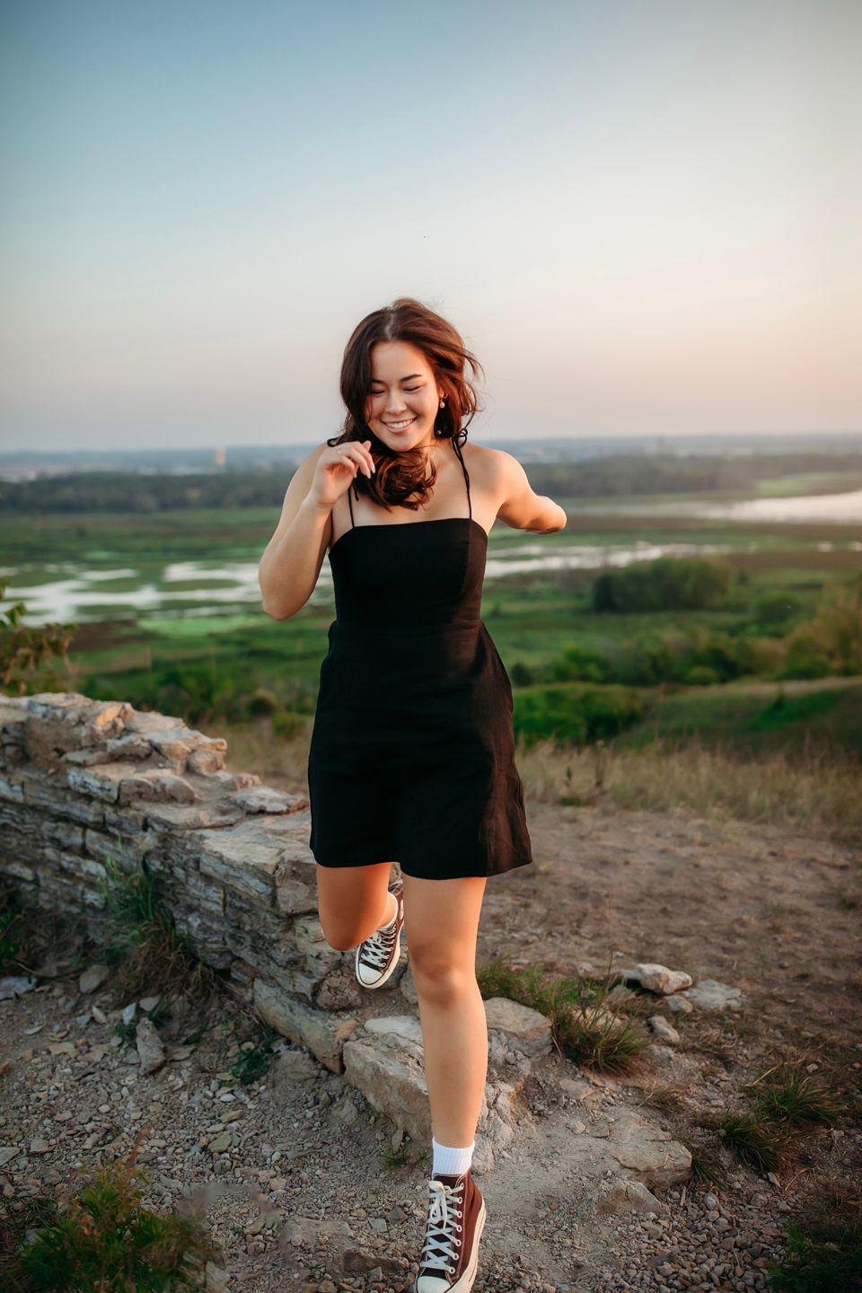 Woman in black dress and sneakers poses playfully on rocky overlook with valley view.
