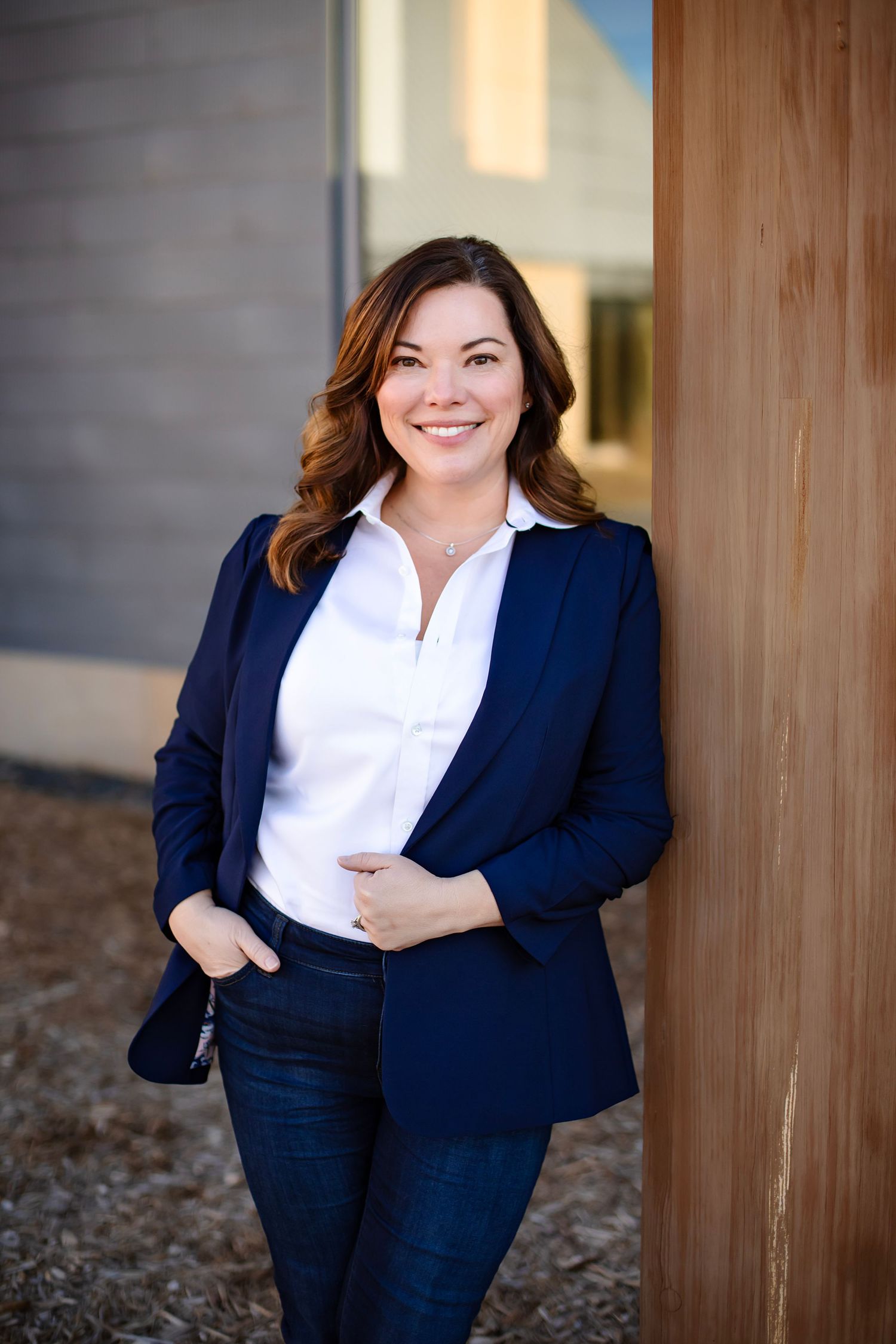 Professional in navy blazer and white blouse poses confidently against wooden wall outdoors.